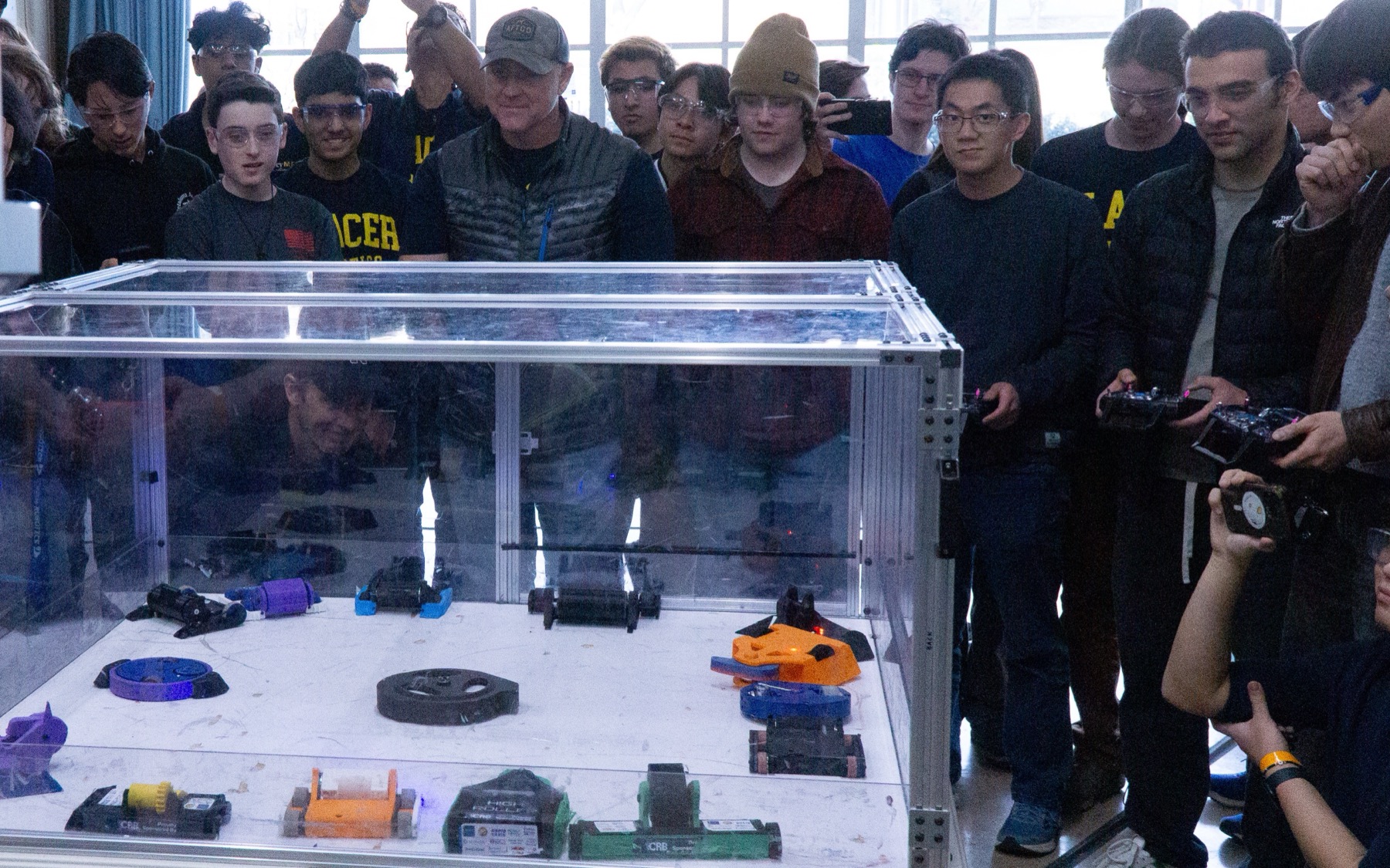 A plastic-ant arena packed with a dozen student-built combat robots mid-rumble, with a crowd of students watching through the polycarbonate